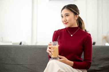 Güzel Asyalı kadın, oturma odasında ya da dinlenme odasında bacak bacak üstüne atmış bir şekilde otururken elinde buzlu kahve tutuyormuş gibi gülümsüyor. Boş zaman, dinlenme, evde dinlenme..