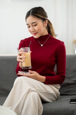 Güzel Asyalı bir kadın oturma odasında ya da dinlenme odasında bacak bacak üstüne atmış bir şekilde otururken buzlu kahve içmekten hoşlanır. Boş zaman, dinlenme, evde dinlenme..