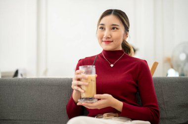 Oturma odasında ya da dinlenme odasında kumaş koltukta otururken gülümseyen ve elinde buzlu kahve tutan Asyalı güzel bir kadın. Boş zaman, dinlenme, evde dinlenme..