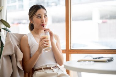 Cam bardakta pipet tutan güzel Asyalı kadın kafenin masasından dışarı bakarken buzlu kahve içiyor. Boş Zaman Rahatlatıcı, Dışarısı Rahatlatıcı.