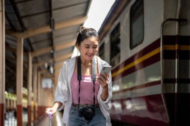 Güzel Asyalı kadın telefona bakıp, tren istasyonunda bavulu bir kenara bırakıp gülümsüyor. Tayland Tren İstasyonu, Turist Taşımacılığı.