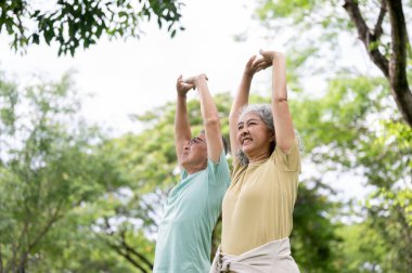 Yaşlı adam ve yaşlı kadın parkta ayakta durup kollarını başlarının üstüne uzatırken gülümsüyorlar. Yaşlı çift, Yeşil Doğa, Serinleyen Açık Hava.