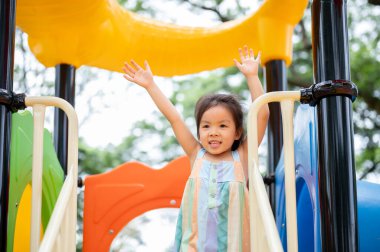 Gülümseyen Asyalı kız parkın oyun parkında renkli bir tırmanma kaydırağında el sallıyor. Yaratıcı Oyuncaklar, sevimli çocuklar, Aile Gezisi.