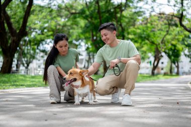 Genç Asyalı gülümseyen bir adam parktaki kaldırımda bir kadınla çömelirken elinde bir Corgi köpeği tasması tutuyor. Çift tişört, evcil köpek, Aile Gezisi..