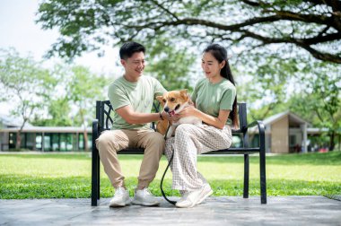Gülümseyen Asyalı erkek ve kadın parkın bankında oturduktan sonra dinleniyorlar. Çift tişört, evcil köpek, Aile Gezisi..