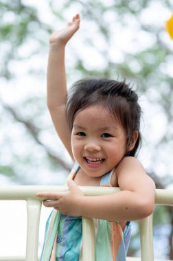 Gülümseyen Asyalı kız, parkın oyun bahçesinde renkli bir tırmanma kaydırağına elini kaldırıyor. Yaratıcı Oyuncaklar, sevimli çocuklar, Aile Gezisi.