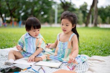 Kız kardeşler kalem tutarak çizim defterine bakarken parkta çim üzerinde piknik paspası üzerinde oturuyorlar. Tatlı Asyalı çocuklar, Aile Gezisi.