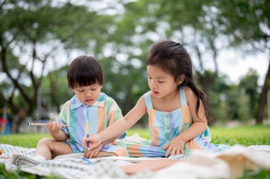 Piknik minderinin üzerine oturmuş parkta çimlerin üzerinde yatarken elinde kalem öğretmenliği yapan kız kardeş defterine çizim yapıyor. Tatlı Asyalı çocuklar, Aile Gezisi.