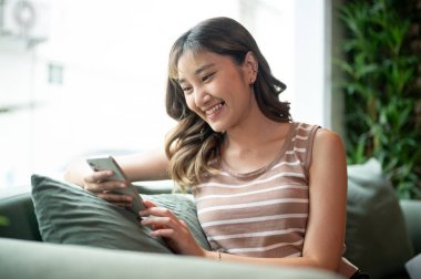 Kafede ya da kantinde yeşil kanepeye yaslanmış otururken telefona bakan gülümseyen Asyalı kadın. Sosyal Ağ, Dışarıdaki Serinleme, Günlük Yaşam.