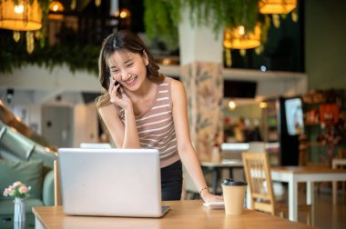 Telefon görüşmesi sırasında gülümseyen Asyalı kadın kafeteryada ya da kantinde dizüstü bilgisayara bakmak için eğildi. Çalış ya da çalış, dışarıda takıl, günlük hayatta..
