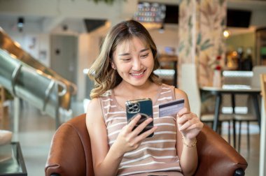 Kahverengi kanepede ya da kantinde oturan Asyalı kadın elinde telefonla kredi kartına bakıyor. Banka reklamları, nakit ödemeler, günlük yaşam..
