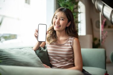 Beyaz ekranlı telefon gösteren Asyalı bir kadın bir kafe veya salonda yeşil kanepeye yaslanmış oturuyor. Haberler ve güncelleme, Reklamcılık Mockup, Günlük yaşam.