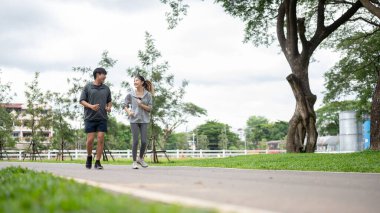 Spor kıyafetli Asyalı kadın ve erkek parkta koşarken ya da koşarken gülümseyip konuşuyorlardı. Genç çift, eşofman. Yeşil Doğa.