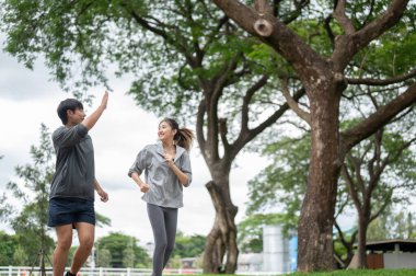 Asyalı bir adam elini kaldırıyor ve spor kıyafetli bir kadın koşarken gülümsüyor ya da parkta kaldırımda koşuyor. Genç çift, eşofman. Yeşil Doğa.