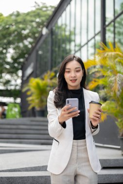 Asyalı kadın ofis çalışanı ya da iş kadını telefona bakıyor ve elinde kahveyle binanın önünde duruyor. Şirket işi, öğle yemeği arası, modern şirket..