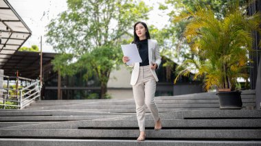 Asyalı kadın ofis çalışanı ya da iş kadını elinde dizüstü bilgisayar ve belgeyle binadan çıkarken. Şirket işi, modern şirket, dışarıda çalışmak..
