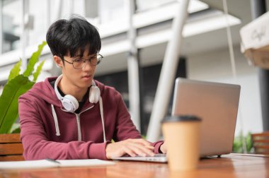 Gözlüklü Asyalı ya da üniversite öğrencisi elinde dizüstü bilgisayarla kafedeki tahta masada kahve içerken daktilo kullanıyor. Çalış ya da çalış, açık hava bahçesi..