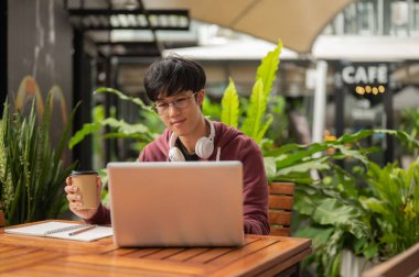 Kafedeki ahşap masada otururken elinde kahve olan Asyalı gözlüklü ya da üniversite öğrencisi dizüstü bilgisayara bakıyor. Çalış ya da çalış, açık hava bahçesi..