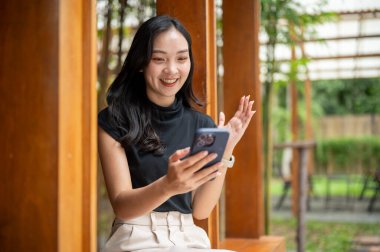 Kafenin ahşap koridorunda bambu bahçesinin yanında otururken Asyalı kadın telefona bakıyor ve elini kaldırıyor. Boş zaman, dışarıda dinlenme zamanı.