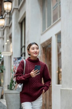 Alışveriş merkezinde ya da markette yürürken elinde telefon tutan örgü ören Asyalı güzel bir kadın. Seyahat turisti, sonbahar giysisi, dışarıda dinlenme..