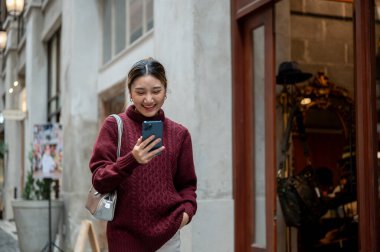 Örgü kazaklı güzel bir Asyalı kadın alışveriş merkezinde ya da markette yürürken telefona bakıyor. Seyahat gezisi, sonbahar giysisi, dışarıda dinlenme..