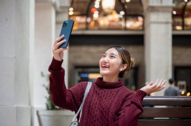 Gülümseyen Asyalı kadın alışveriş merkezindeki bankta otururken telefon çekiyor ya da video çekiyor. Seyahat gezisi, sonbahar giysisi, dışarıda dinlenme..