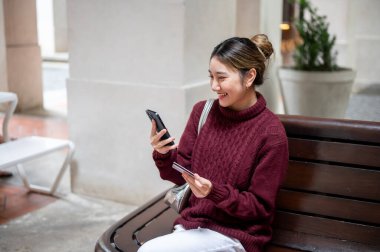 Elinde kredi kartı olan gülümseyen Asyalı kadın alışveriş merkezinde bankta otururken telefona bakıyor. Seyahat parası, Nakitsiz Toplum, Banka reklamları.