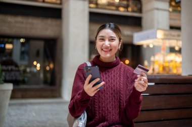 Alışveriş merkezinde ya da markette bankta otururken elinde kredi kartı ve akıllı telefon tutan Asyalı bir kadın. Seyahat parası, Nakitsiz Toplum, Banka reklamları.