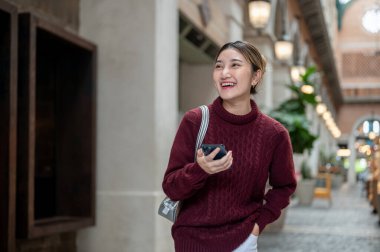 Kırmızı kazaklı gülümseyen Asyalı kadın alışveriş merkezinde ya da markette yürürken telefonunu kaçırıyor. Seyahat turisti, sonbahar giysisi, dışarıda dinlenme..