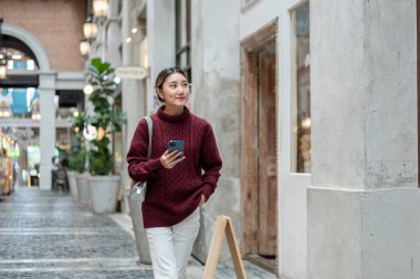 Örgü kazak giyen güzel bir Asyalı kadın elinde telefonla alışveriş merkezinde ya da markette geziniyor. Seyahat turisti, sonbahar giysisi, dışarıda dinlenme..