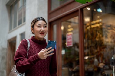 Örgü ören süveter giyen gülümseyen Asyalı kadın elinde telefonla alışveriş merkezinde ya da markette geziniyor. Seyahat gezisi, sonbahar giysisi, dışarıda dinlenme..