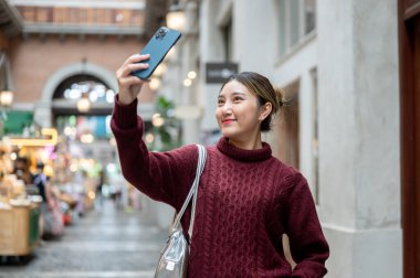 Örgü kazaklı güzel bir kadın elinde telefonla alışveriş merkezinde ya da markette fotoğraf çekiyor. Seyahat gezisi, sonbahar giysisi, dışarıda dinlenme..