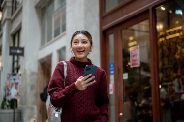 Örgü süveter giyen güzel bir Asyalı kadın elinde telefon, ayakta ya da alışveriş merkezinde ya da markette yürüyor. Seyahat gezisi, sonbahar giysisi, dışarıda dinlenme..