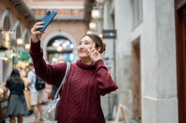 Örgü kazaklı Asyalı kadın elinde telefonla alışveriş merkezinde ya da markette Ziyaretçi barışı iç çekerken fotoğraf çekiyor. Seyahat gezisi, sonbahar giysisi, dışarıda dinlenme..