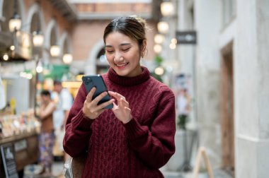 Örgü ören süveter giymiş gülümseyen Asyalı bir kadın alışveriş merkezinde ya da markette telefonuna bakıyor. Seyahat gezisi, sonbahar giysisi, dışarıda dinlenme..
