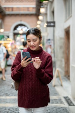 Alışveriş merkezinde ya da markette yürürken telefonda yazan örgü ören Asyalı genç bir kadın. Seyahat turisti, sonbahar giysisi, dışarıda dinlenme..