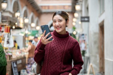 Örgü ören süveter giymiş gülümseyen Asyalı bir kadın elinde telefonla bir alışveriş merkezinde ya da markette yürüyordu. Seyahat gezisi, sonbahar giysisi, dışarıda dinlenme..