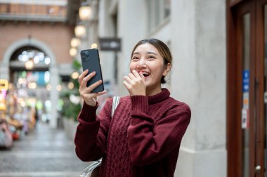 Bir alışveriş merkezinde ya da markette elinde telefonla kahkaha atan örgü ören Asyalı bir kadın. Seyahat turisti, sonbahar giysisi, dışarıda dinlenme..