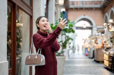 Örgü ören kazaklı, elinde telefonla alışveriş merkezinde ya da markette fotoğraf çeken Asyalı bir kadın. Seyahat gezisi, sonbahar giysisi, dışarıda dinlenme..