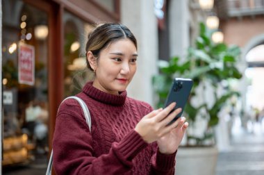Örgü kazaklı genç bir kadın elinde telefonla alışveriş merkezinde ya da markette çekilmiş fotoğraflara bakıyor. Seyahat turisti, sonbahar giysisi, dışarıda dinlenme..