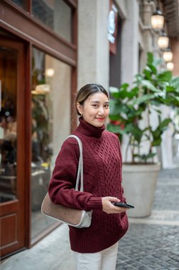 Örgü kazaklı genç Asyalı bir kadın alışveriş merkezi ya da marketin koridorunda ayakta ve elinde telefonla bekliyor. Seyahat turisti, sonbahar giysisi, dışarıda dinlenme..