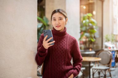 Genç Asyalı kadın alışveriş merkezinde ya da markette ayakta dikilip akıllı telefon tutarak kazak örüyor. Seyahat turisti, sonbahar giysisi, dışarıda dinlenme..