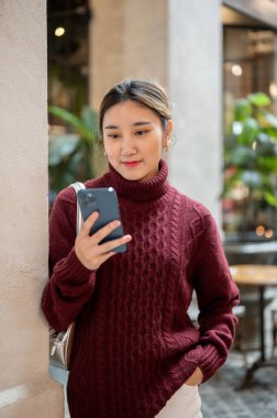 Örgü süveter giyen Asyalı bir kadın alışveriş merkezinde ya da markette cep telefonuna bakıyor. Seyahat gezisi, sonbahar giysisi, dışarıda dinlenme..