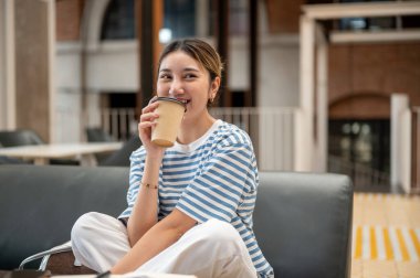 Gülümseyen Asyalı genç bir kadın kafede deri koltukta bacak bacak üstüne atarak kahve içmekten zevk alır. Boş zaman, dışarıda dinlenme, şehir hayatı..