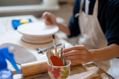 Seramik bardaktan resim fırçası alan bir çömlekçinin eli sınıftaki ya da stüdyodaki çalışma masasında. Boyama, El yapımı Ürün, Sanat Atölyesi.