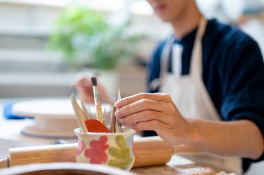Genç çömlek sanatçısı seramik bardaktan boya fırçasını çalışma masasında ya da stüdyoda alıyor. Çizim veya Renklendirme, El yapımı Ürün, Sanat Atölyesi.