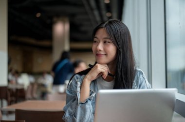 Kot ceketli genç Asyalı kadın elinde kalemle kahvede dizüstü bilgisayarından kafasını çeviriyor. Çalışmak ya da Çalışmak, Serbest Çalışmak, Şehir Hayatı.