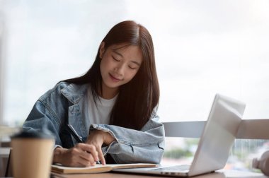 Kot ceketli genç Asyalı kadın, kafedeki ahşap masaya dizüstü bilgisayarla birlikte not defterine yazıyor. Çalışmak ya da Çalışmak, Serbest Çalışmak, Şehir Hayatı.