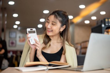 Kafedeki tahta masada otururken elinde kalem ve hesap makinesi olan gülümseyen Asyalı kadın dizüstü bilgisayara gösteriyor. Çalışmak ya da çalışmak, şehir hayatı, dışarıda dinlenmek..