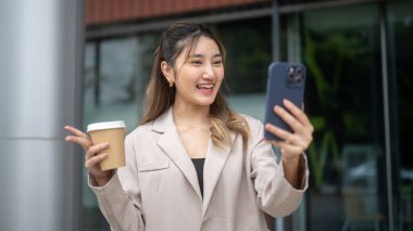 Elinde kahve tutan Asyalı bir kadın bir kafe önünde dikilirken telefonla fotoğraf ya da görüntülü konuşma yapıyor. İletişim, Şehir hayatı, Dışarıdaki Serinleme.
